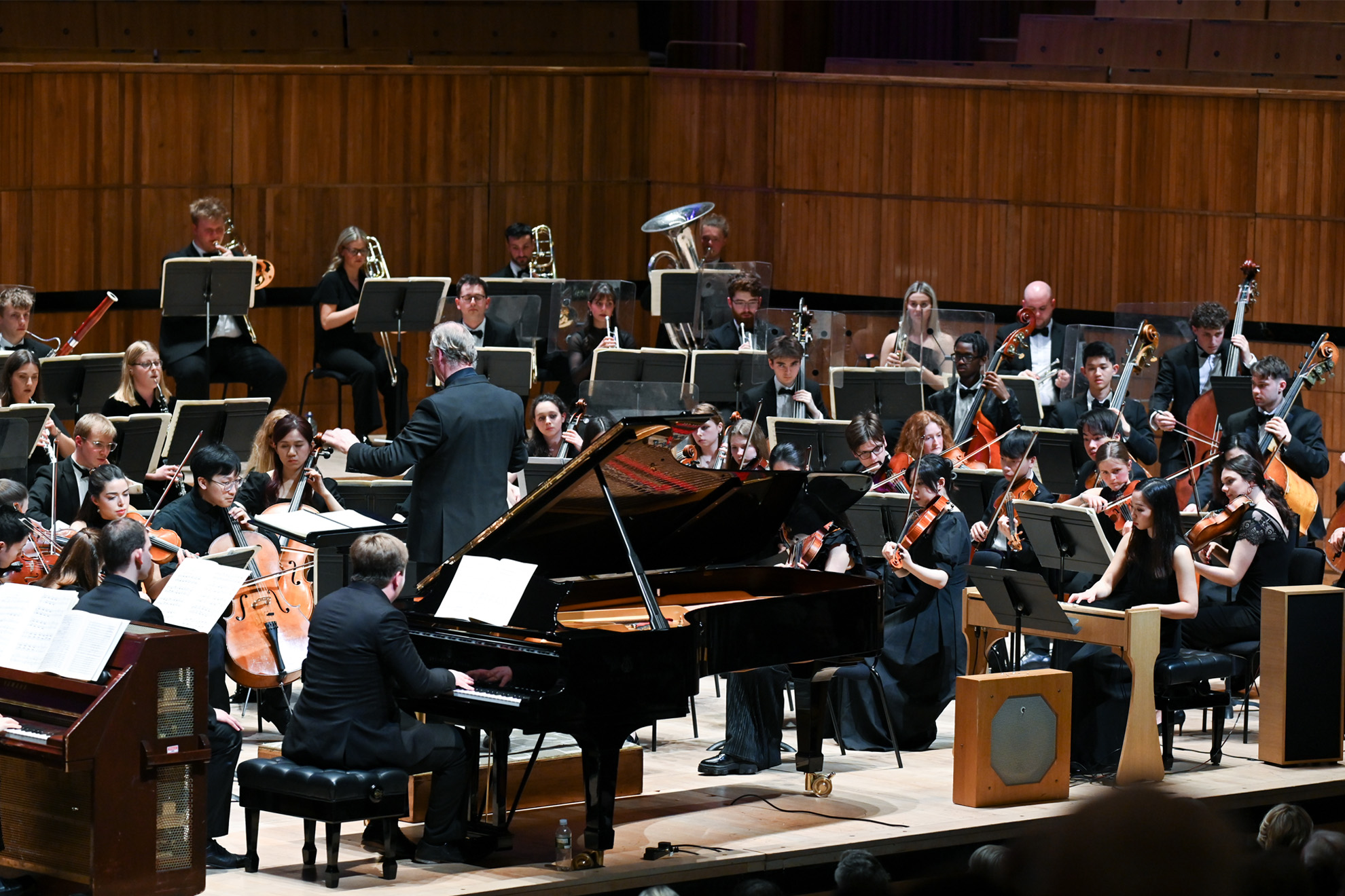 An full orchestra performance, wearing smart black attire, with multiple pianos and ondes Martenot near the front of the stage, performing to an audience.